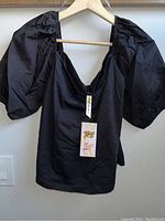 Front view of the black women's top on a hanger showing puffy sleeves and square neckline with attached brand tags.
