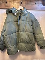 Front view of green puffer jacket on hanger showing hood with drawstrings, quilted design and button detail on collar.
