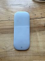 Top view of a white Microsoft Surface Mouse on a wooden surface, showing the slim and smooth top with a small Microsoft logo.