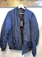 Medium blue bomber jacket hanging on white hanger shows front side with zipper and ribbed cuffs and waistband.