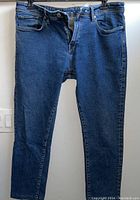 Front full view of blue denim jeans hanging on black hanger, showing classic five-pocket style and button-fly closure.