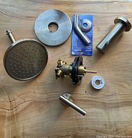 Overview of all shower set components laid on wood surface: stainless steel showerhead, brass valve assembly, spout, flange plate, plumbing tape, and mounting parts.