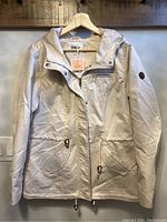 Front view of the women's canvas parka hanging on a wooden hanger, showing button and zipper closure, front pockets, and brown patch on sleeve.