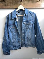 Front view of medium blue denim jacket hanging on wooden hanger showing button front and chest pockets