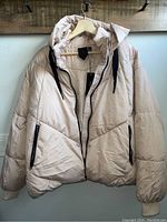 Front view of beige puffer jacket hanging on hanger, showing full front with hood and zipper pockets.