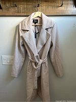 Full view of light beige women's coat hanging, showing lapel collar and belt tie.