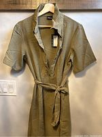 Front view of olive green XL shirt dress with a belt tied around the waist, hanging on a wooden hanger on a wall.