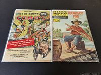 Photo shows two vintage comic books face up on a dark surface. Left comic is Smilin' Ed's Buster Brown Comics No. 15 with colorful cover, right is Classics Illustrated No. 19 'Huckleberry Finn' with illustrated fishing scene cover.
