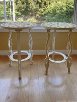 Two white cast iron tables with decorative cut-out tops and tripod decorative legs, standing on wood floor near window