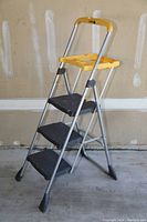Photo of the 3-step folding ladder with yellow tool tray, metallic frame, and black steps, standing upright against a wall.