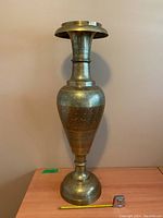 Full view of the tall brass vase or stand showing its overall shape and size relative to a measuring tape on the table below.