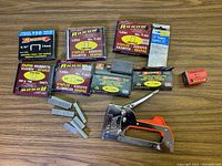 Overall view of Arrow Fastener staple gun and multiple boxes of Arrow T50 staples including 3/8 inch and 1/2 inch sizes with some loose staples laid out.