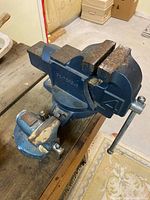 Photo showing two bench vises mounted on wooden plank, one larger blue vise in back and smaller blue vise with brass jaw plates in front.