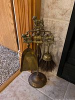 Full view of brass fireplace tool set standing on floor near fireplace with shovel, brush, poker, and stand with dog figurines on top and tool handles.