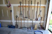 Wide view of gardening tools lined up against garage wall, showing shovels, rakes, broom, and other tools