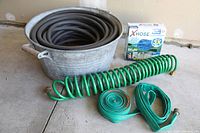 Photo showing large galvanized metal tub with a coiled black hose inside, green spiral hose, flat green hoses, and boxed expandable hose on concrete floor.