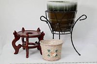 Wooden plant pot stand, ceramic pot in black iron stand, and small woven basket with floral design and plastic lining arranged on white surface with measuring scale in front