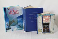 Photo of three hardcover sailing books placed standing side by side. From left to right: The Illustrated Encyclopedia of World Sailing with a dust jacket featuring a sailboat, Piloting Seamanship & Small Boat Handling in solid blue hardcover, and Small Craft Piloting & Coastal Navigation with a patterned cover including a compass image.
