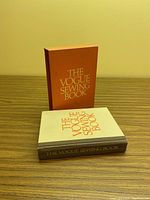Shows the hardcover book with white dust jacket titled 'The Vogue Sewing Book' alongside the orange protective sleeve/box with the title in silver text.