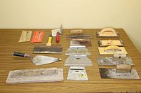 Overview of various trowels and sanding tools arranged on a wooden table, showing different sizes and shapes of tools with wooden handles and metal blades.