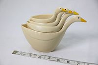 Four cream-colored plastic nesting measuring cups shaped like geese with yellow beaks, shown stacked inside each other on a white surface next to a measuring tape for size reference.