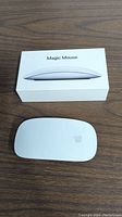 Apple Magic Mouse placed on a surface next to its original box showing product side profile image and name.