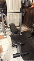 Weight bench with metallic frame, black padded seat and leg attachment, barbell rack with barbell and weight plates attached.