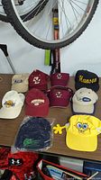 Table with a total of 10 various caps and hats including maroon sports caps, white Lakers cap, navy Under Armour cap in plastic, and yellow SpongeBob cap.