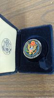 Round metal military pin in blue velvet lined case, showing vibrant colors and text.