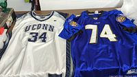 Photo showing two sports shirts lying flat, white UConn basketball jersey No. 34 and blue Baltimore Ravens football jersey No. 74.