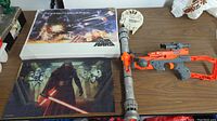Photo showing two completed Star Wars themed jigsaw puzzles and one orange and gray toy blaster gun on a table.