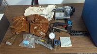 Full lot overview on table showing various hand tools, leather tool belt, pneumatic nailer, dial indicator, and tote bag.