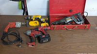 Top view of five power tools on a table including several cordless drills and a reciprocating saw in a red metal case.