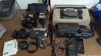 Photo of all items together on a table, showing camera, flash units, vintage camcorder, battery charger, cables, and beige carrying case.