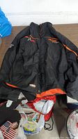 Front view of black racing jacket with orange accents laid flat on table, showing embroidered logos and full length zipper.