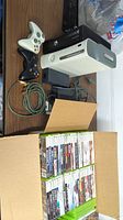 Photo of 2 Xbox 360 consoles: white and black, with 2 controllers placed next to them and a box containing multiple games.
