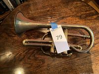Brass trumpet laying horizontally on wooden surface, showing bell and valve assembly, with auction tag attached.
