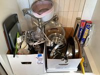 Two boxes filled with kitchen cookware and utensils including Calphalon stainless steel pots and pans, baking sheets, mixing bowls, and kitchen utensils.