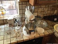 White KitchenAid stand mixer with attached stainless steel bowl, additional stainless bowl on top, white ceramic bowl and stack of clear glass mixing bowls on tiled counter