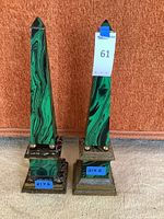 Two of the green and black marble obelisk statues with stepped metal bases shown against an orange backdrop