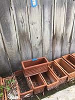 Six rectangular terracotta planter boxes arranged on ground against wood fence, one stacked