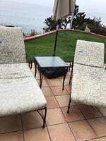 Chaise lounges and part of patio table with umbrella base visible on tiled patio outdoors with greenery background.