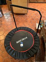 Top view of round black trampoline with red trim and KanChiMi logo, including attached padded black handlebar.
