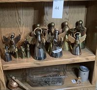 Wood shelf with multiple brass metal angel candle holders of different sizes, placed next to a container with small metal chess pieces and a candle extinguisher.