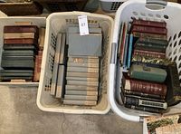Three laundry baskets filled with hardcover books, including grey cloth volumes and assorted cloth-bound titles