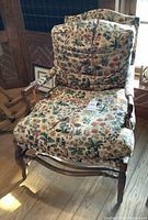 Front view of wood arm chair with floral cushioned seat, backrest, and armrests. Cushion fabric has multicolor floral pattern. Chair positioned near window.