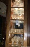 Oak display cabinet with clear glass shelves showing set including clear glass pitcher and punch bowl on top shelf and rows of etched gold rimmed glasses on lower shelves