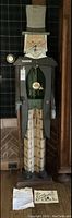 Full front view of painted wooden clock in the shape of a standing gentleman wearing a top hat with pendulum disc visible
