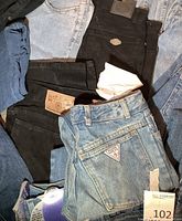 Closer photo showing Guess jeans blue denim with triangular logo patch and other blue and black jeans with brand waistband patches visible.