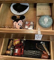 Ceramic plate, Mickey-shaped items, clear plexiglass piece, sealed steak thermometer pack, red marble cutter, black humidor pouch and tobacco pipe in top drawer
